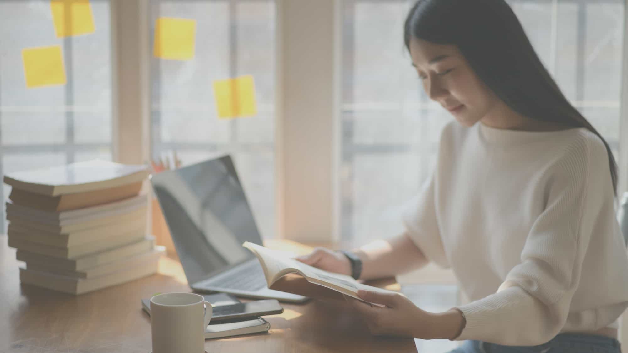 A Woman Reading A Book On A Desk With Laptop, Mug, Books, Phone